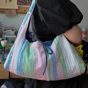 Può includere: Una grande borsa tote colorata realizzata con camicie riciclate. La borsa presenta un design patchwork con strisce verticali e pannelli in rosa pastello, blu, verde e bianco. Ha un interno in denim e una chiusura con bottoni. Il cinturino è realizzato con lo stesso materiale della camicia.