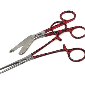 May include: Two stainless steel surgical instruments with red handles featuring a black paw print pattern. The instruments are labeled "Aimée".