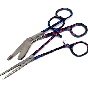 May include: Three surgical instruments with paw print designs. The instruments are a pair of hemostats, a pair of scissors, and a pair of Adson forceps. The hemostats and scissors have black handles with blue and pink paw prints. The Adson forceps have a silver handle with the name "Kristena" engraved on it.