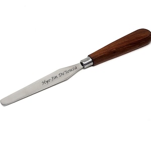 May include: A stainless steel palette knife with a wooden handle. The knife is engraved with the name "Hye-Jin De'Souza".