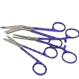 May include: Set of three surgical scissors with purple paw print designs. The scissors are labeled "Name" on the handles.