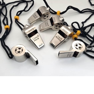May include: A collection of silver-colored soccer coach whistles with black lanyards and yellow beads. Each whistle is engraved with "2023" and a coach's name, along with a soccer ball design. The whistles are displayed on a white surface.