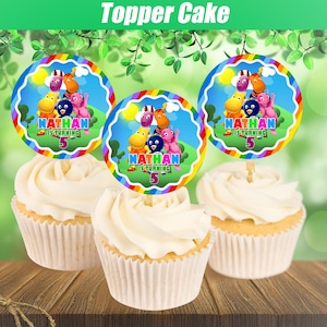 May include: Three cupcakes with white frosting and decorative toppers. The toppers are round with a rainbow border and feature cartoon characters and the text "Nathan is turning 5". The background is a blurred green.