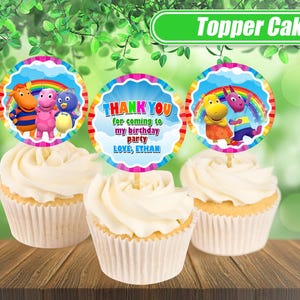 May include: Three cupcakes with white frosting and decorative toppers. The toppers feature cartoon characters, rainbows, and the text "THANK YOU for coming to my birthday party LOVE, ETHAN". The image is set against a blurred green background.