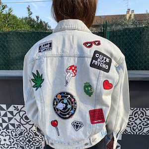 Op de afbeelding: Een lichtblauw denim jack met verschillende patches, waaronder een paddenstoel, een hart, een diamant, een planeet, een marihuanablad, een kroon, een zonnebril en tekst die "Cute but Psycho" en "Empowered Women Empower Women" zegt.