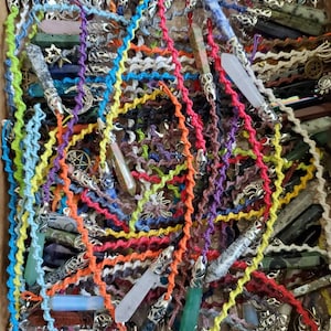 May include: A close-up of a variety of colorful crystal point pendants on knotted cords. The cords are in a variety of colors, including red, orange, yellow, green, blue, purple, and white. The crystal points are also in a variety of colors, including clear, pink, green, blue, and purple.