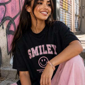 Puede incluir: Una camiseta negra de manga corta con la palabra SMILEY en letras rosas sobre un gráfico de cara sonriente rosa y el texto ORIGINALS EST. 1972. La prenda se combina con pantalones de color rosa claro y una pulsera con pequeños dijes de caritas.