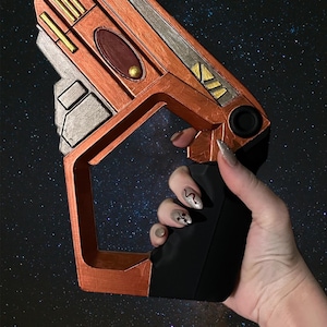 Op de afbeelding: Een hand houdt een futuristisch ogende, oranje en zilveren, handwapen vast met een zwarte grip. Het wapen heeft een rechthoekige vorm met een afgeronde bovenkant en een kleine, zwarte, ronde knop aan de zijkant. De achtergrond is donkerblauw met witte sterren.