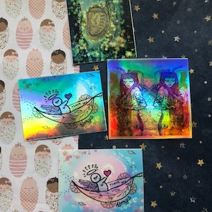 May include: Four iridescent holographic stickers with different designs. One sticker features a drawing of a baby in a womb. Another sticker features a drawing of two angels with wings. The third sticker features a drawing of an angel in a hammock with the text "love you always". The fourth sticker features a drawing of an angel in a hammock with the text "love you always".
