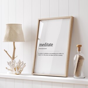 May include: A framed vertical wall art print featuring the dictionary definition of the word meditate in black text on a white background, displayed in a light wood frame alongside a table lamp with a branch base and a glass bottle with a cork.