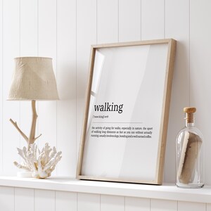 May include: A framed wall art print featuring the dictionary definition of the word walking in black text on a white background. The print is displayed in a light wood frame next to a lamp with a textured shade and a clear glass bottle with a cork.
