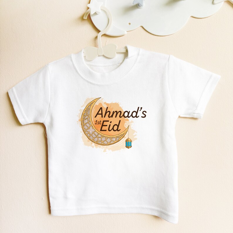 May include: White baby t-shirt with a gold crescent moon and lantern design. The text "Ahmad's 1st Eid" is printed on the shirt.