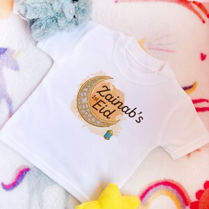 May include: White baby t-shirt with a gold crescent moon and lantern design. The text "Zainab's 1st Eid" is printed on the shirt.