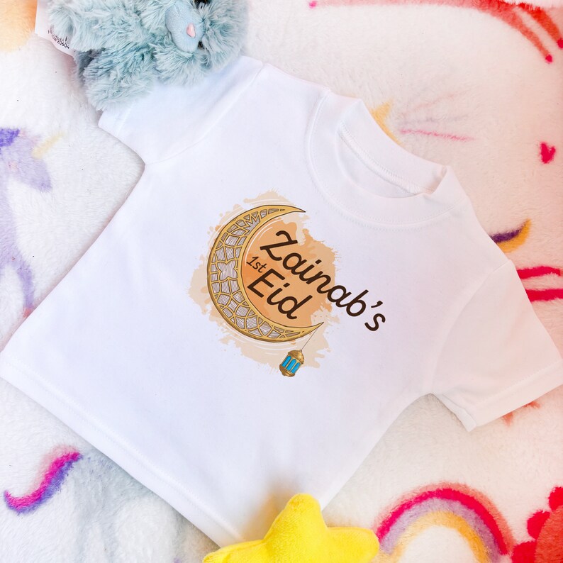 May include: White baby t-shirt with a gold crescent moon and lantern design. The text "Zainab's 1st Eid" is printed on the shirt.