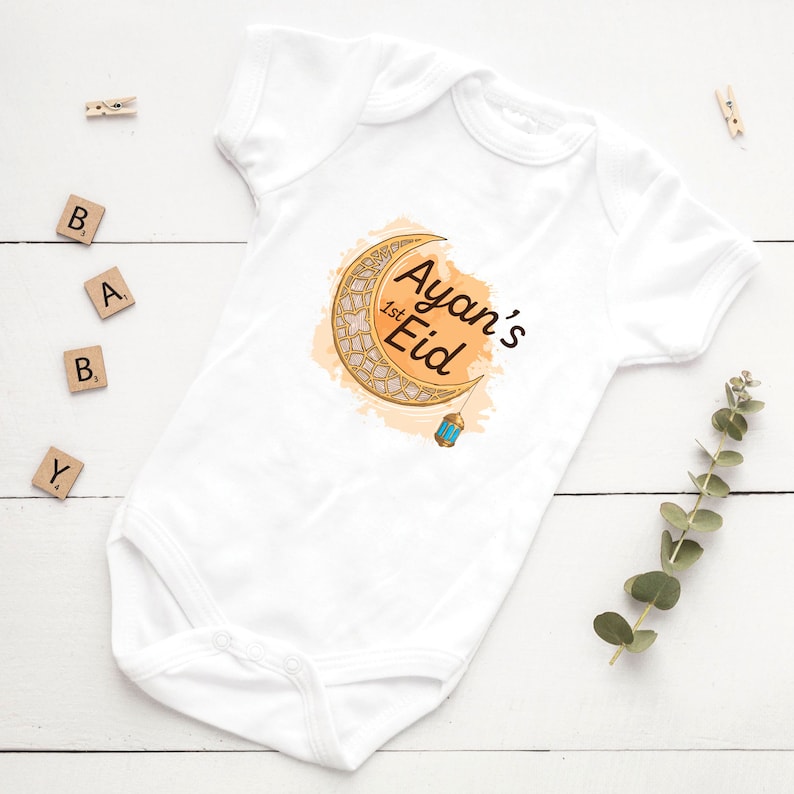 May include: White baby onesie with a gold crescent moon and lantern design. The text "Ayan's 1st Eid" is printed on the onesie.
