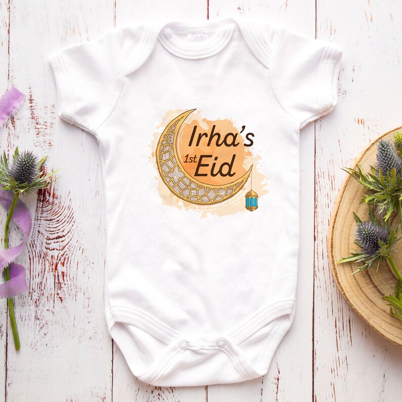 May include: White baby onesie with a gold crescent moon and lantern design. The text "Irha's 1st Eid" is printed on the onesie.