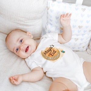 May include: A white baby onesie with a gold crescent moon and the text "Maya's 1st Eid" in brown lettering. The onesie is worn by a baby lying on a white and brown striped blanket.