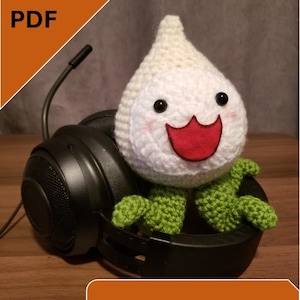 May include: A white crocheted ghost character with green arms and legs sitting on top of a black gaming headset. The ghost has a red smiling mouth and black eyes. The text "PATTERN PDF" and "Instant Download" are visible in the image.