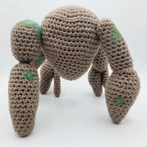 May include: A handmade crochet creature in shades of brown, with a rounded body and multiple leg-like appendages. The plush toy has accents of green and blue on its body. The soft sculpture is displayed against a white background.