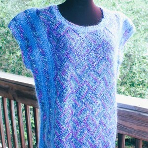 May include: A sleeveless, blue knit top with a round neckline. The top features a textured pattern with shades of blue and purple. The garment is displayed on a black mannequin against a blurred outdoor background.