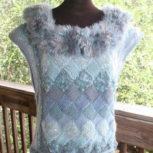 May include: A sleeveless, hand-knitted top in shades of blue and gray, displayed on a mannequin. The top features a textured, diamond-patterned design and a fluffy, faux fur collar. The bottom hem has a decorative border.