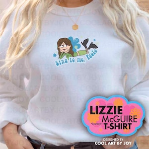 May include: White sweatshirt featuring a cartoon illustration of a character with the text "Sing to me, Paolo." A pink and blue cloud-shaped graphic below reads "Lizzie McGuire T-Shirt" and "Inspired." Designed by Cool Art by Joy.