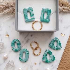 Può includere: Una collezione di gioielli composta da due orecchini rettangolari, due ovali e due triangolari in resina marmorizzata verde e bianca, insieme a tre orecchini a cerchio in metallo color oro disposti su una superficie piana.