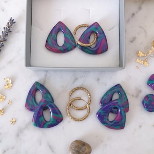 Peut inclure: Un ensemble de boucles d'oreilles triangulaires présentant un motif marbré dans des tons de violet, bleu et vert. Le lot comprend deux paires de pièces triangulaires, deux paires de créoles texturées dorées et deux petits clous ronds.