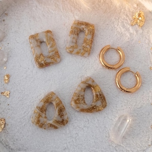 May include: A set of jewelry featuring two rectangular and two triangular translucent white polymer clay charms with embedded gold leaf flakes, accompanied by a pair of plain gold-toned metal hoop earrings, all arranged on a textured surface.