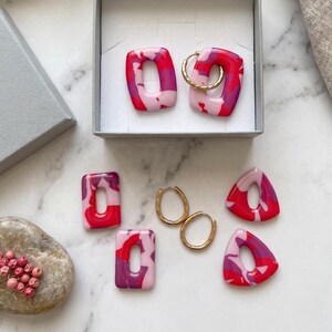 Peut inclure: Une collection de boucles d'oreilles aux formes rectangulaires et triangulaires avec un motif marbré dans des tons rouge, magenta et rose clair. L'ensemble comprend deux paires de créoles en métal doré et plusieurs breloques en argile polymère.