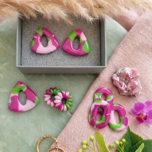 Könnte beinhalten: Eine Schmuckkollektion bestehend aus zwei Paar dreieckigen Ohrringen mit einem marmorierten Muster in Pink, Magenta und Grün, einem Paar kleiner spiralförmiger Ohrringe sowie einem Paar goldfarbener Creolen mit einer strukturierten Oberfläche.