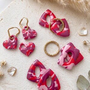 May include: A collection of handmade polymer clay earrings in shades of red, pink, and white. The earrings feature heart, triangle, and rectangular shapes, some with gold-colored hoops. The marbled effect adds a unique touch to each piece.