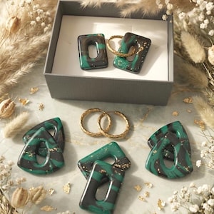 May include: A collection of jewelry pieces, including earrings and hoops, presented in a gray box and on a marble surface. The earrings feature a green and black marbled design with gold accents. Gold hoop earrings are also displayed.