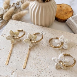 Könnte beinhalten: Goldene Creolen-Ohrringe mit weißen Blumenanhängern und goldenen Stäben, und ein Paar goldene Creolen-Ohrringe mit weißen Blumenanhängern. Die Ohrringe werden auf einer gesprenkelten, hellen Oberfläche präsentiert.