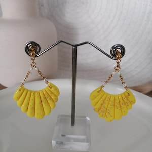 以下が含まれることがあります： 手作りの黄色のイヤリングのペア。扇形のデザインで、ポリマークレイ製。ゴールドカラーのアクセントと繊細なチェーンが特徴です。イヤリングはシルバーのイヤリングスタンドに展示されています。