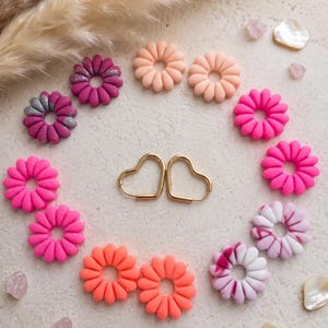 Könnte beinhalten: Eine Sammlung farbenfroher, blütenförmiger Ohrringe und Accessoires. Die Ohrringe sind um ein Paar goldfarbene herzförmige Creolen angeordnet. Die Blumen sind in Rosa-, Orange- und Pfirsichtönen gehalten, einige mit Glitzerakzenten. Dekorative Elemente sind Kristalle und Muscheln.