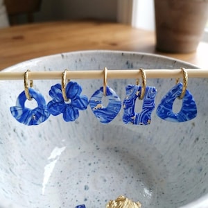 May include: A collection of five blue and white marbled polymer clay earrings with gold-colored accents. Each earring hangs from a gold hoop. The earrings are displayed on a wooden dowel inside a speckled ceramic bowl.