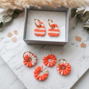 May include: A collection of earrings featuring two teardrop-shaped pieces with horizontal orange and white stripes on gold hoops, and three circular, fluted flower-shaped pieces in a marbled orange and white pattern, two of which are on gold hoops.