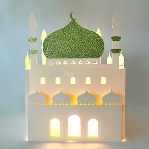 Könnte beinhalten: Ein weißes Papiermodell einer Moschee mit einer grünen Glitzerkuppel und beleuchteten Fenstern. Die Moschee hat mehrere Minarette und Bögen.