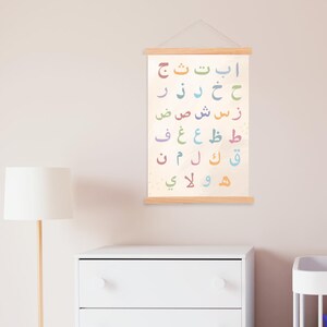 Könnte beinhalten: Eine farbenfrohe arabische Alphabettabelle mit hellbeigem Hintergrund. Die Buchstaben sind in verschiedenen Farben gehalten, darunter Orange, Grün und Blau. Die Tabelle wird von Holzstäben gehalten und an einer Schnur aufgehängt. Die Tabelle wird an einer hellrosa Wand angezeigt.