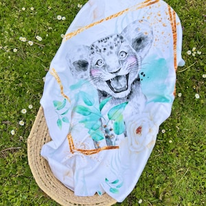 Puede incluir: Manta de bebé blanca con un diseño de acuarela que presenta un cachorro de tigre blanco con hojas verdes y una rosa blanca. La manta tiene un borde geométrico dorado.