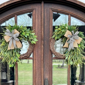 May include: Two green and brown wreaths with black and white striped bows hang on a wooden double door with glass panels.