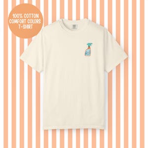 May include: Cream-colored t-shirt with a small graphic of a spray bottle on the front. The shirt has the text "100% Cotton Comfort Colors T-Shirt" in a circular orange background. The shirt is made of a soft, comfortable material.