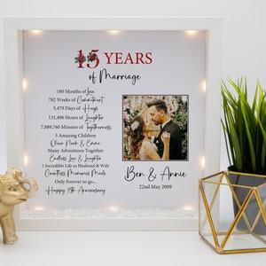 May include: A white light-up picture frame with a photo of a couple and a red and green floral design. The frame has a white background with text that reads "15 YEARS of Marriage" and lists the number of months, weeks, days, hours, and minutes they have been married. The text also includes the names of their children, "Oliver, Noah & Evie", and a message that reads "Happy 15th Anniversary". The couple's names, "Ben & Annie", and their wedding date, "22nd May 2009", are printed below the photo.