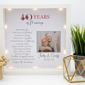 May include: A white light-up frame with a photo of a couple celebrating their 40th anniversary. The frame has a red and white 40 with a floral design and the text "40 Years of Marriage". The text also includes the number of months, weeks, days, hours, and minutes they have been together. The couple's names are Judy & Geoff and the date is 17th May 1984.
