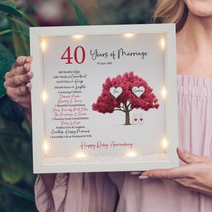 May include: A framed print celebrating a 40th wedding anniversary. The white frame contains a design featuring a red tree, two heart-shaped name tags, and the text "Happy Ruby Anniversary." Small lights illuminate the frame.