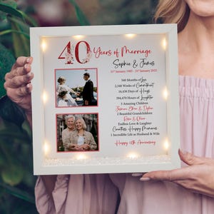 May include: A white light-up frame with two photographs and a message celebrating 40 years of marriage. The message includes the couple's names, Sophie and James, and the dates 31 January 1985 - 31 January 2025. The message also includes a list of milestones, such as 540 months of love, 2,348 weeks of commitment, 16,436 days of hugs, 394,470 hours of laughter, 3 amazing children, 2 beautiful grandchildren, endless love and laughter, countless happy memories, and 1 incredible life as husband and wife. The frame is decorated with a red 40 with floral accents.