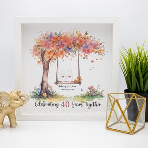 May include: A white framed picture with a watercolor painting of a tree with pink and orange flowers. Two figures are sitting on a swing hanging from the tree. The text "Aubrey & Victor 14th February 1981 Celebrating 40 Years Together" is printed below the image.