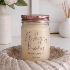 Può includere: Candela in barattolo di vetro con coperchio color rame, etichettata "Blueberry Pancakes" in corsivo. La candela è posta in un piatto di pietra decorativo. L'etichetta include anche "WILLOW LAYNE CO." e "340g/12oz."
