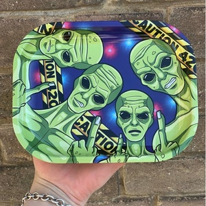 Puede incluir: Bandeja rectangular de metal con cuatro figuras de alienígenas verdes mostrando el dedo medio. La bandeja tiene un fondo azul con explosiones de estrellas y cinta amarilla de precaución con el texto "CAUTION 42".